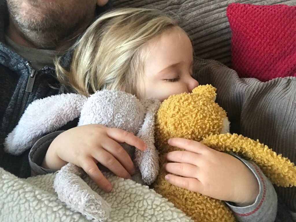 A young child resting with her father clutching her cuddly toys in her hands.